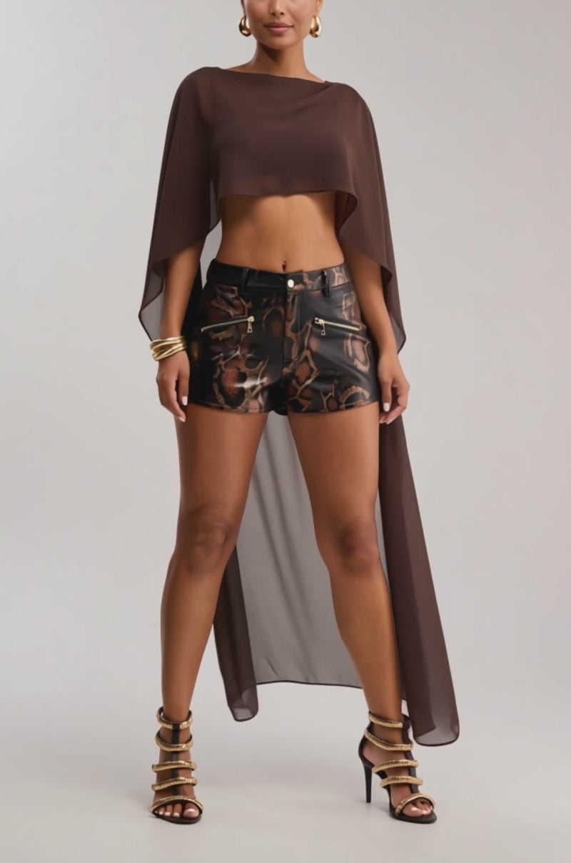 Video of woman modeling the IT GIRL BLOUSE IN BROWN, a sheer chiffon high-low top with a flowing cape, paired with black metallic high-waisted shorts, gold jewelry, and strappy heels against a simple light background.