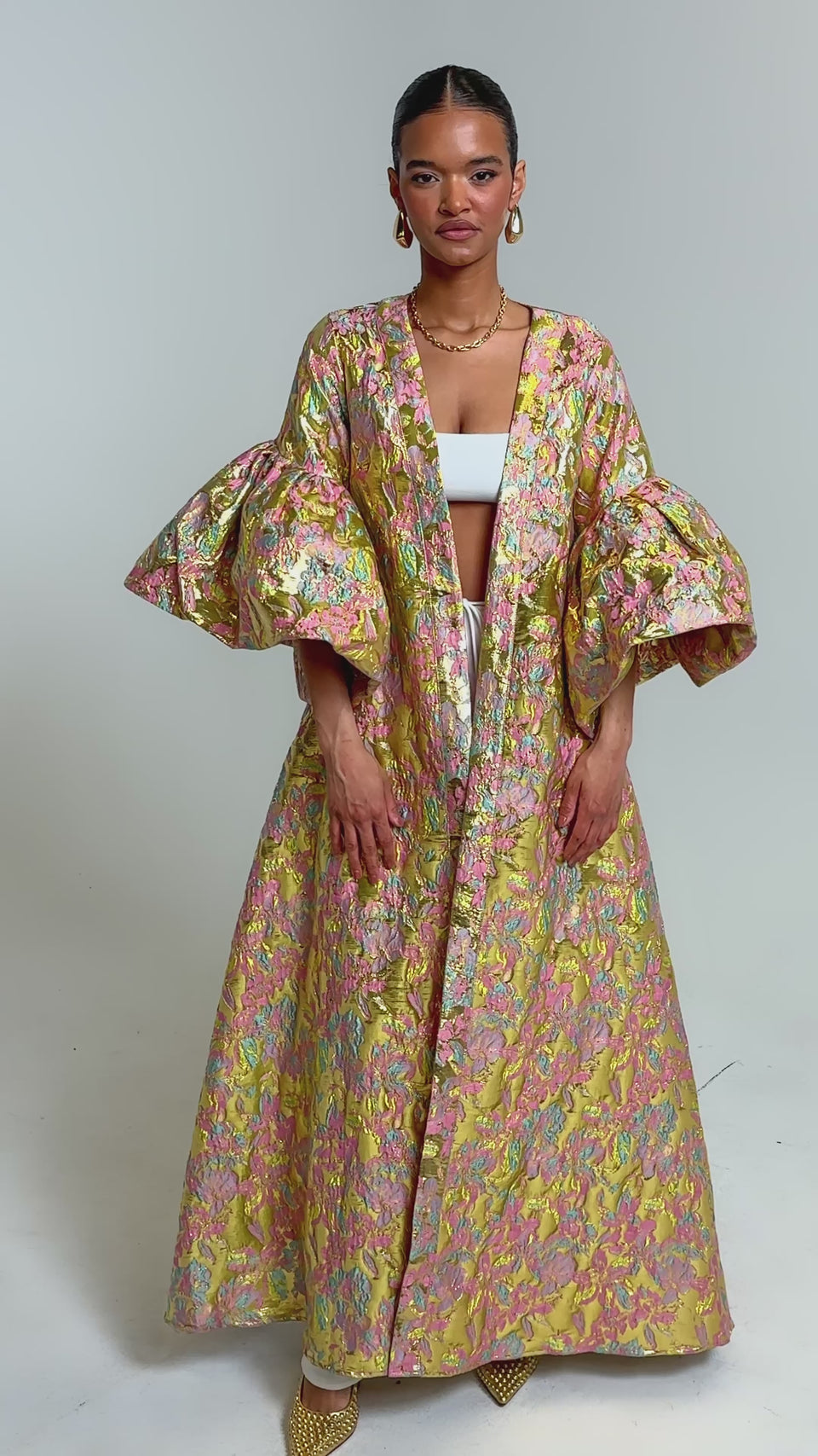 Video of woman modeling the LEMON DROP BROCADE DUSTER, featuring metallic pink, gold, and green hues with voluminous sleeves. She pairs it with a pastel top, white pants, gold earrings, and pointed shoes for a standout look.
