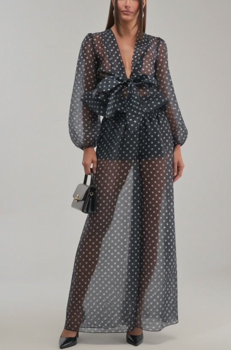 A video of a woman in the PRETTY GIRL POLKA DOT BLOUSE—a sheer black top with white dots, long puffed sleeves, deep V-neck, and a large waist bow.