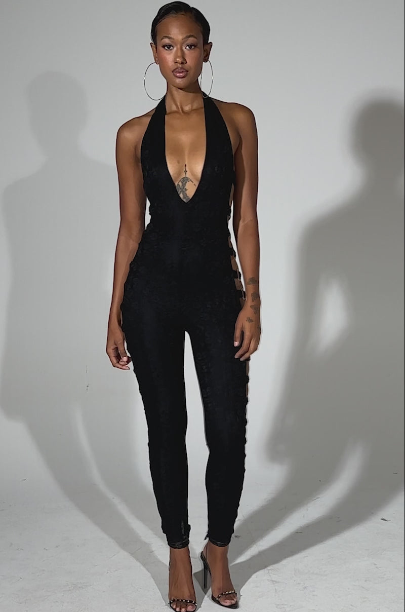 A video of a woman modeling the NOT FOR EVERYONE CUTOUT LACE JUMPSUIT, featuring black lace fabric, bold side cutouts with straps, and paired with high heels against a plain light background.