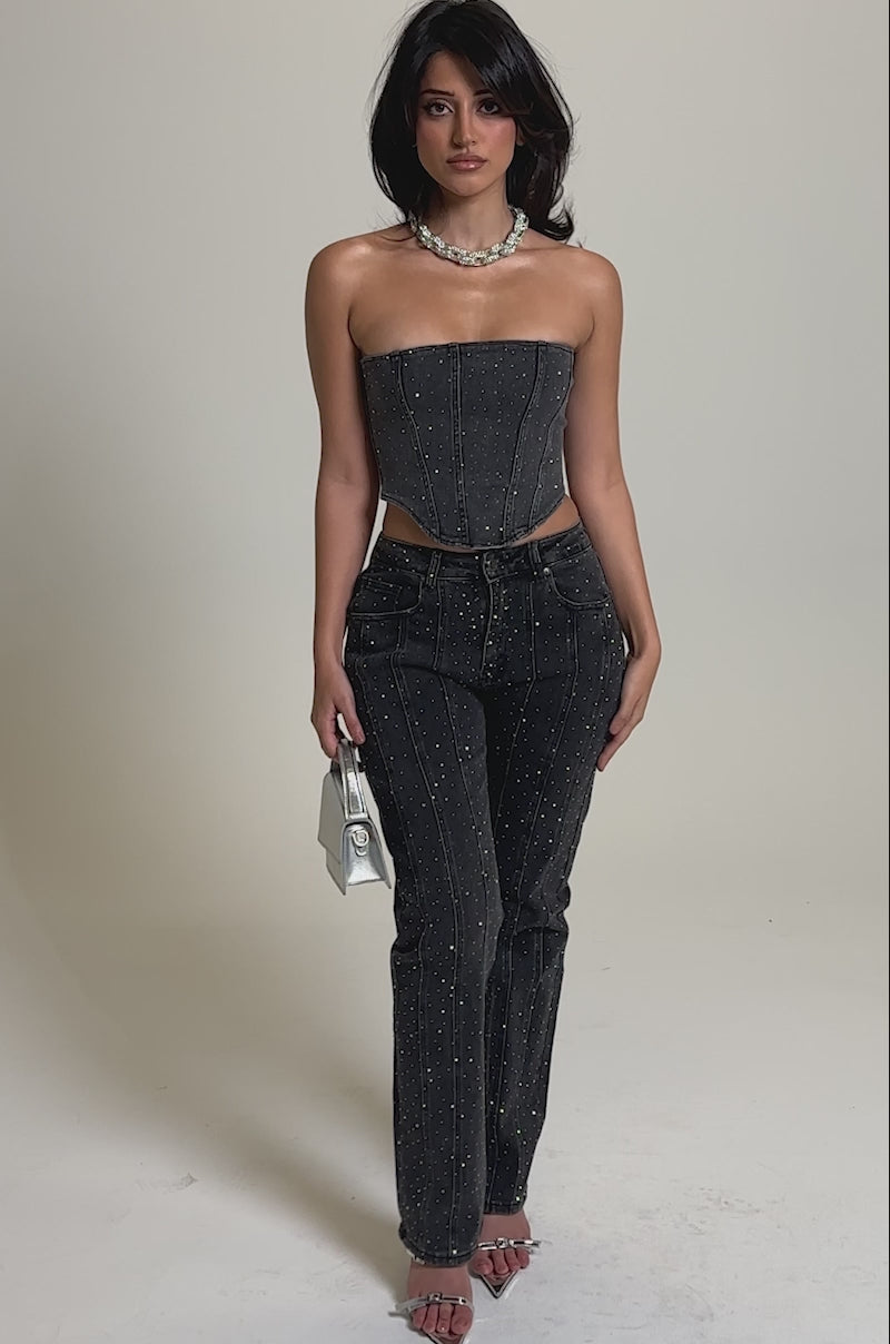 A video of a woman wearing a strapless gray top and LETS GET IT RELAXED DENIM PANT with sparkling rhinestone details, silver heels, a chunky necklace, and holding a small silver handbag.