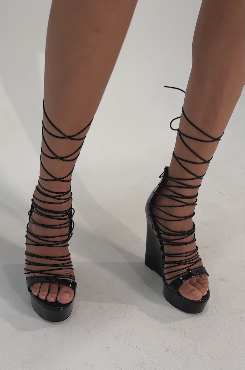 A video of a model wearing the AKIRA GOLD TAVI BLACK SWEDGE SANDAL WITH LACE UP.