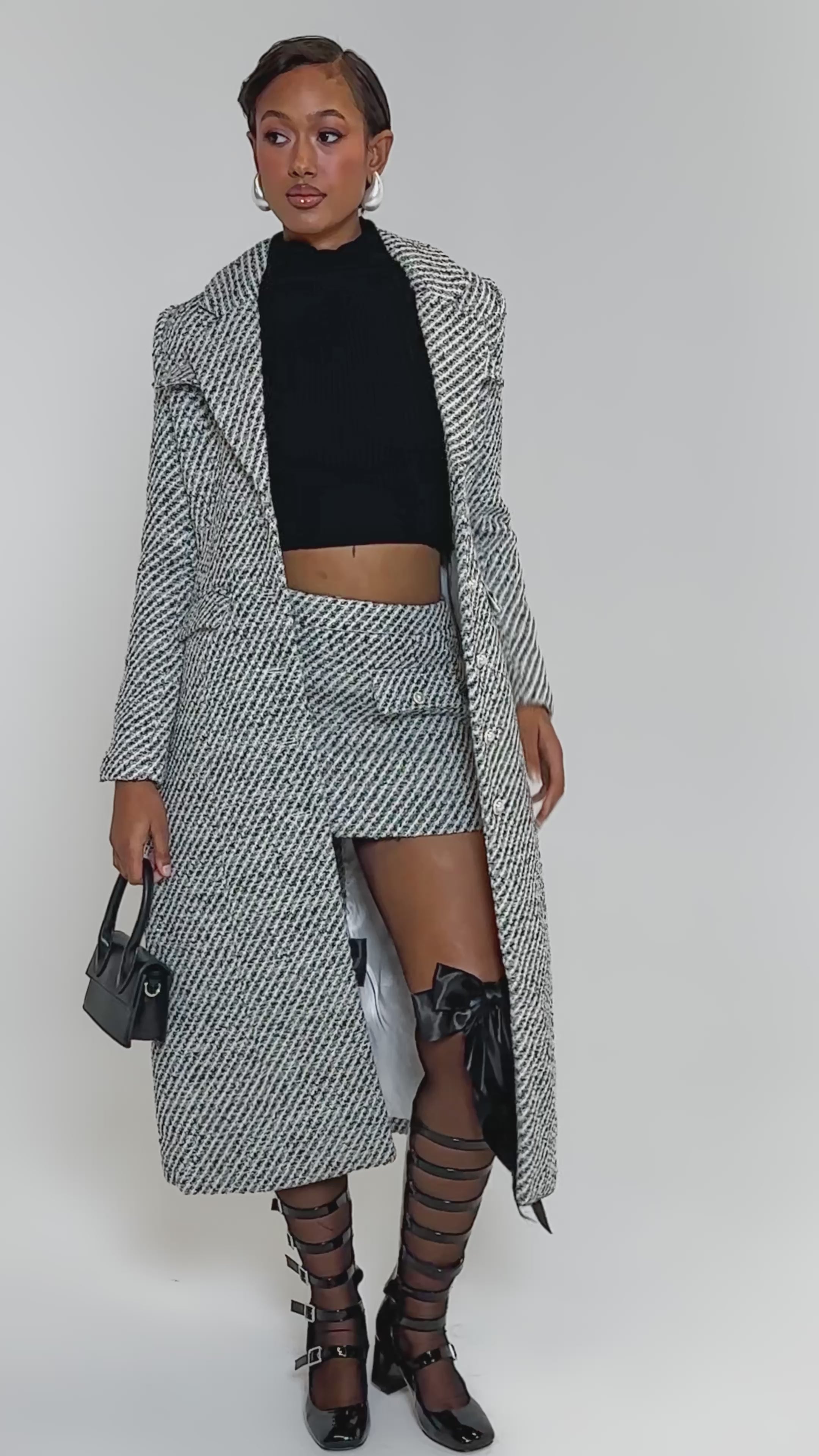 Video of woman modeling the SEVILLE TWEED TRENCH, paired with a matching mini skirt, black crop top, knee-high black strappy boots, and a small black handbag against a plain white background highlighting her winter elegance.