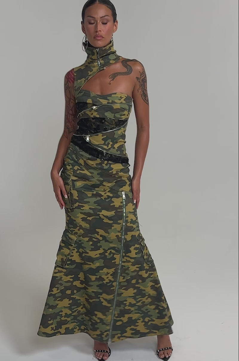 Video of woman modeling the FUSED TOGETHER MOCK NECK MAXI DRESS IN CAMO, styled with a black belt, hoop earrings, and black heeled sandals against a plain white background.