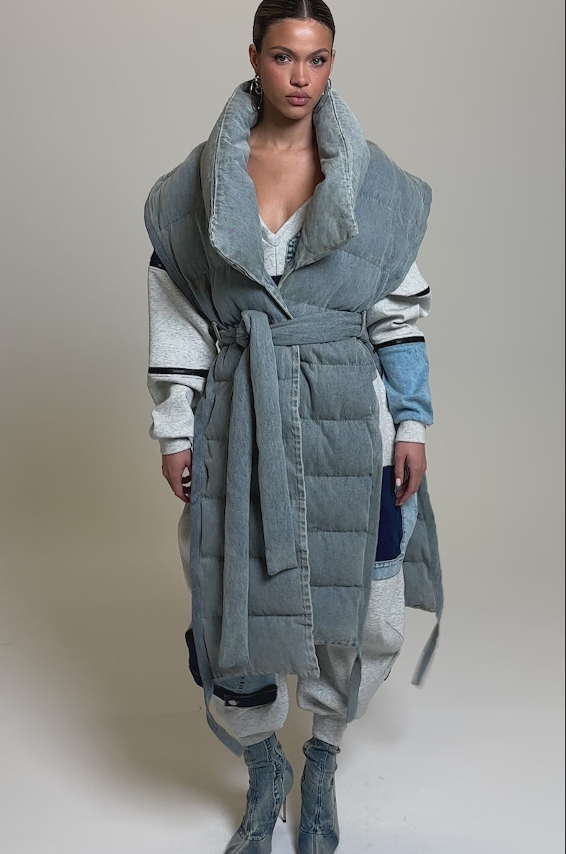 Video of woman modeling the PILOT JONES LONG LINE PUFFER VEST IN DENIM, belted at the waist over a gray tracksuit and blue boots, against a plain light background.