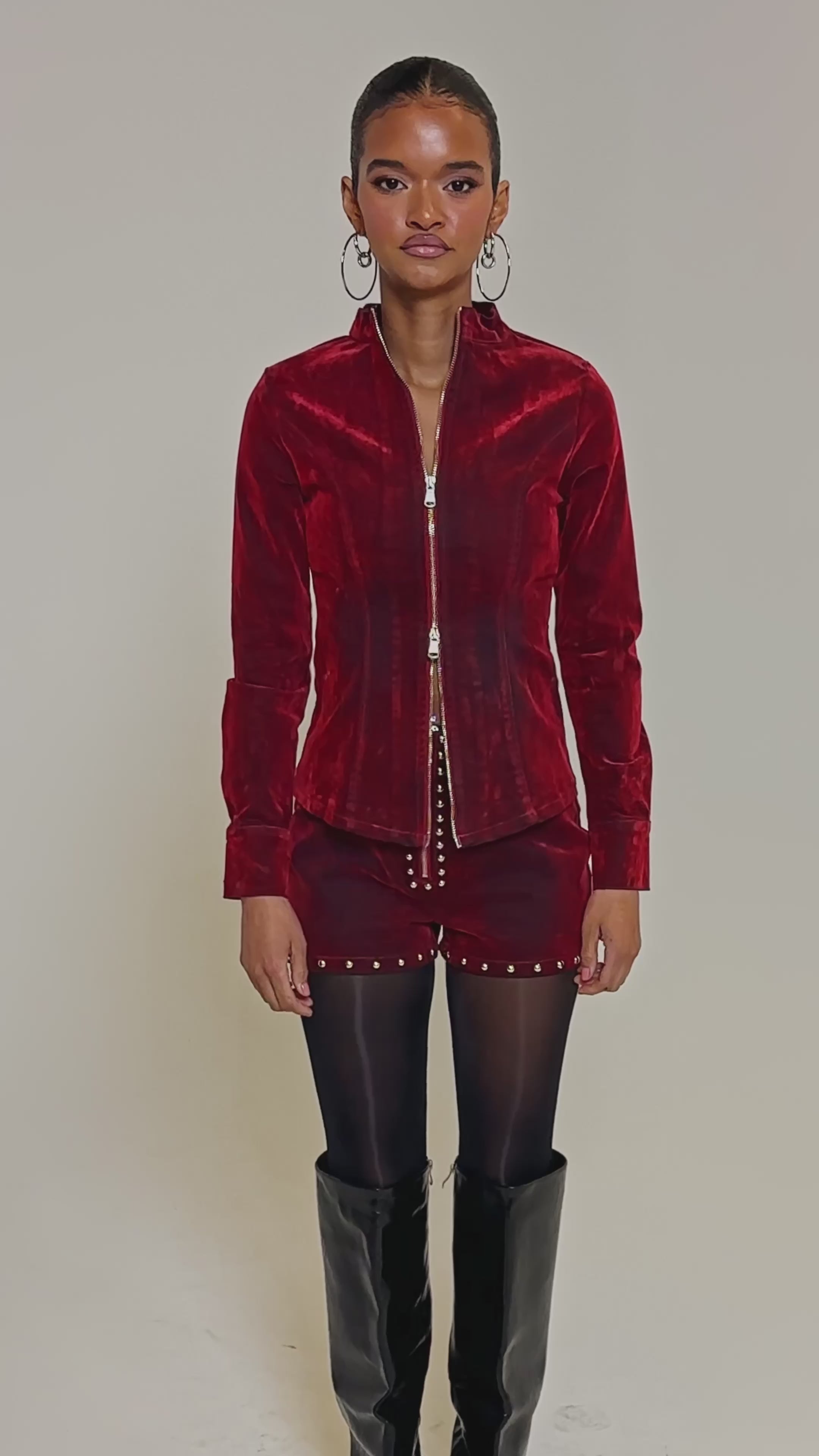 Video of woman modeling the RODEO VIBES VELVET DENIM ZIP UP jacket with matching red studded shorts, accessorized with hoop earrings and dark tights, channeling a modern cowgirl look against a plain light background.
