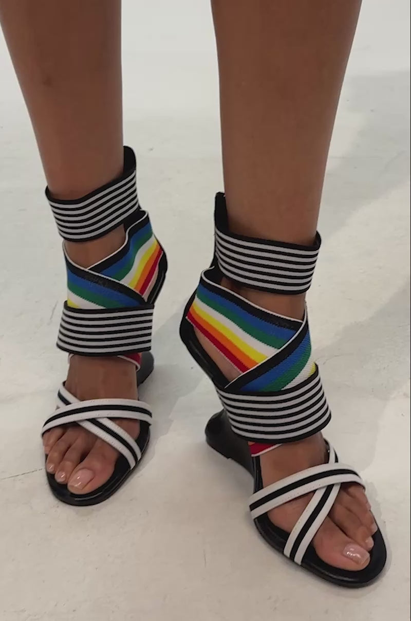 Video of woman modeling the AKIRA GOLD SIGMA MULTI WEDGE SANDAL, which features black inverted wedge heels, white straps, multicolored rainbow elastic bands, black and white striped ankle straps, and a comfort cushion insole, set against a plain background.