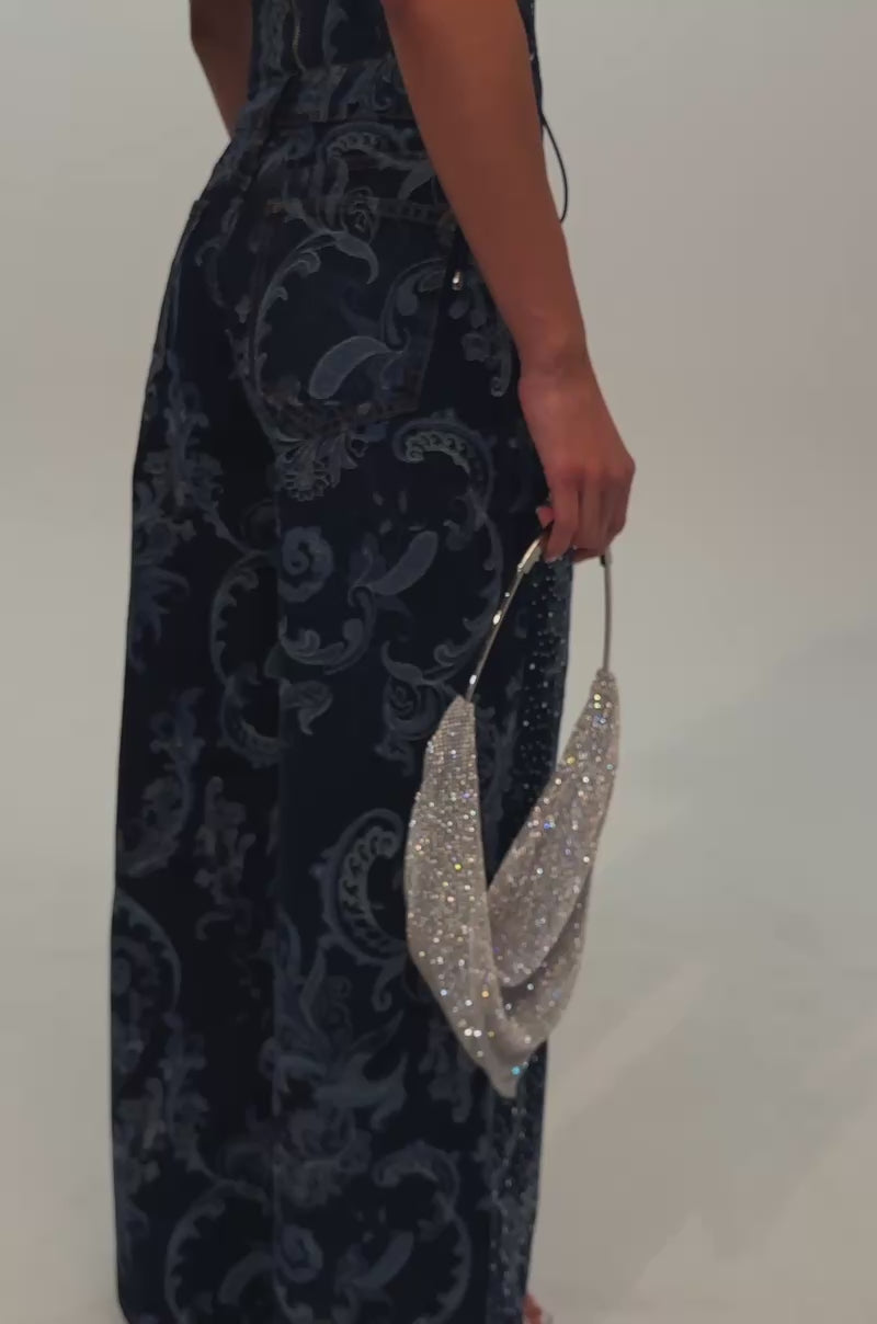 A video of a woman wearing a strapless blue paisley corset top and the ELEVATED WIDE LEG DENIM WITH RHINESTONES, which features a flattering high-rise waist, holds a silver bag while standing against a plain light background.
