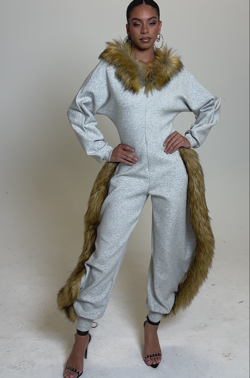 A video of a woman modelling the YOU WISH JUMPSUIT WITH FUR ACCENTS, featuring bold brown faux fur around the collar, hips, and ankles. She pairs it with black high-heeled sandals against a simple light background.