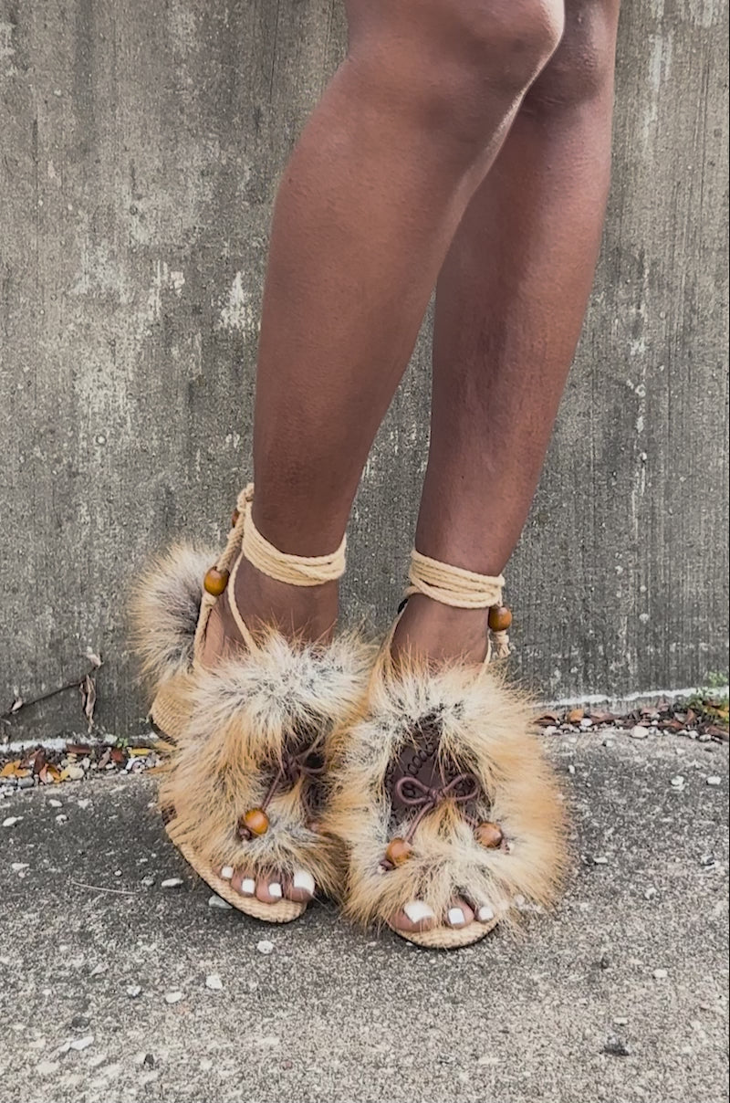 Video of woman modeling the AKIRA GOLD UMIKA BROWN FUR CHUNKY SANDAL, featuring faux fur and bead details, while standing outdoors against a concrete wall.
