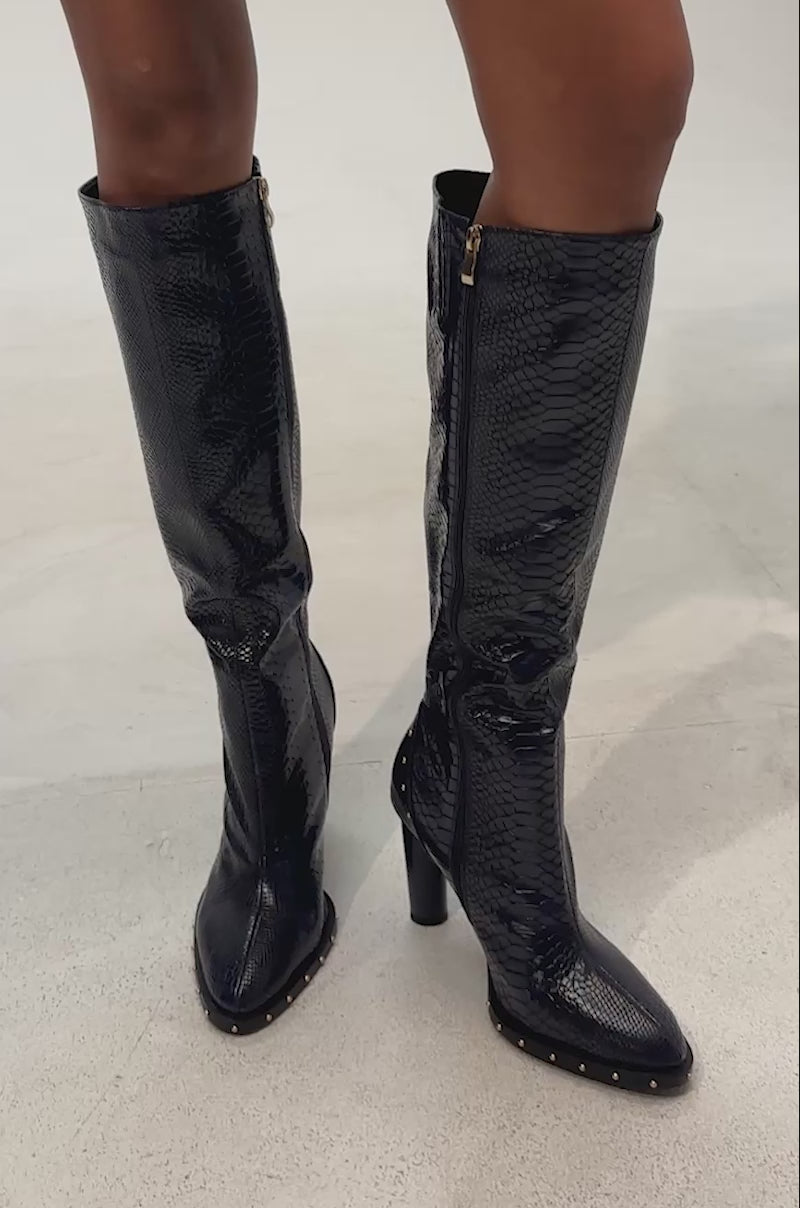 Video of woman modeling the AZALEA WANG LIOREN NAVY SNAKE BLOCK KNEE HIGH BOOT- a single, dark blue snake-print boot with a high block heel and metallic studs along the sole near the toe