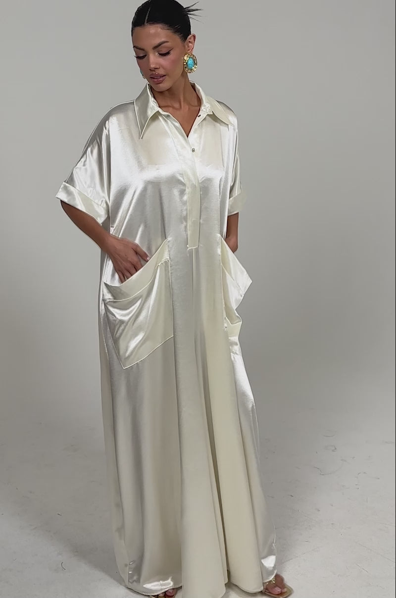 Video of woman modeling the SHE'S RICH OVERSIZED JUMPSUIT IN LUXE CREAM, featuring a collared neckline and short sleeves, paired with bold turquoise earrings against a plain white background.