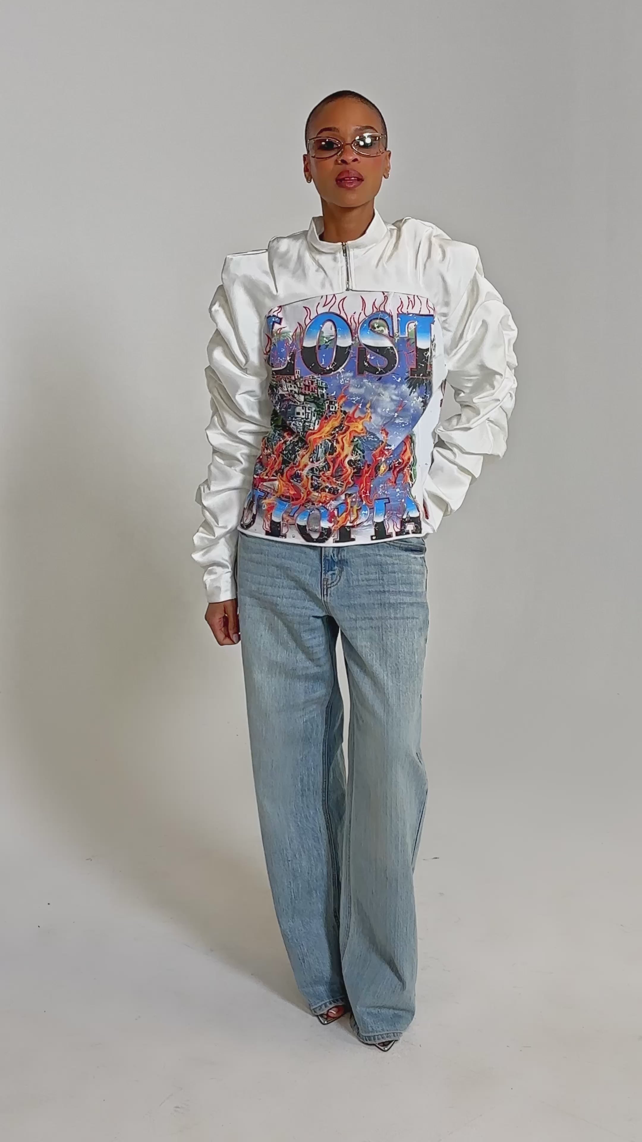 Video of woman modeling the CORA SATIN BOLERO IN WHITE featuring ruched sleeves and a vibrant "LOST UTOPIA" graphic of flames and city imagery.