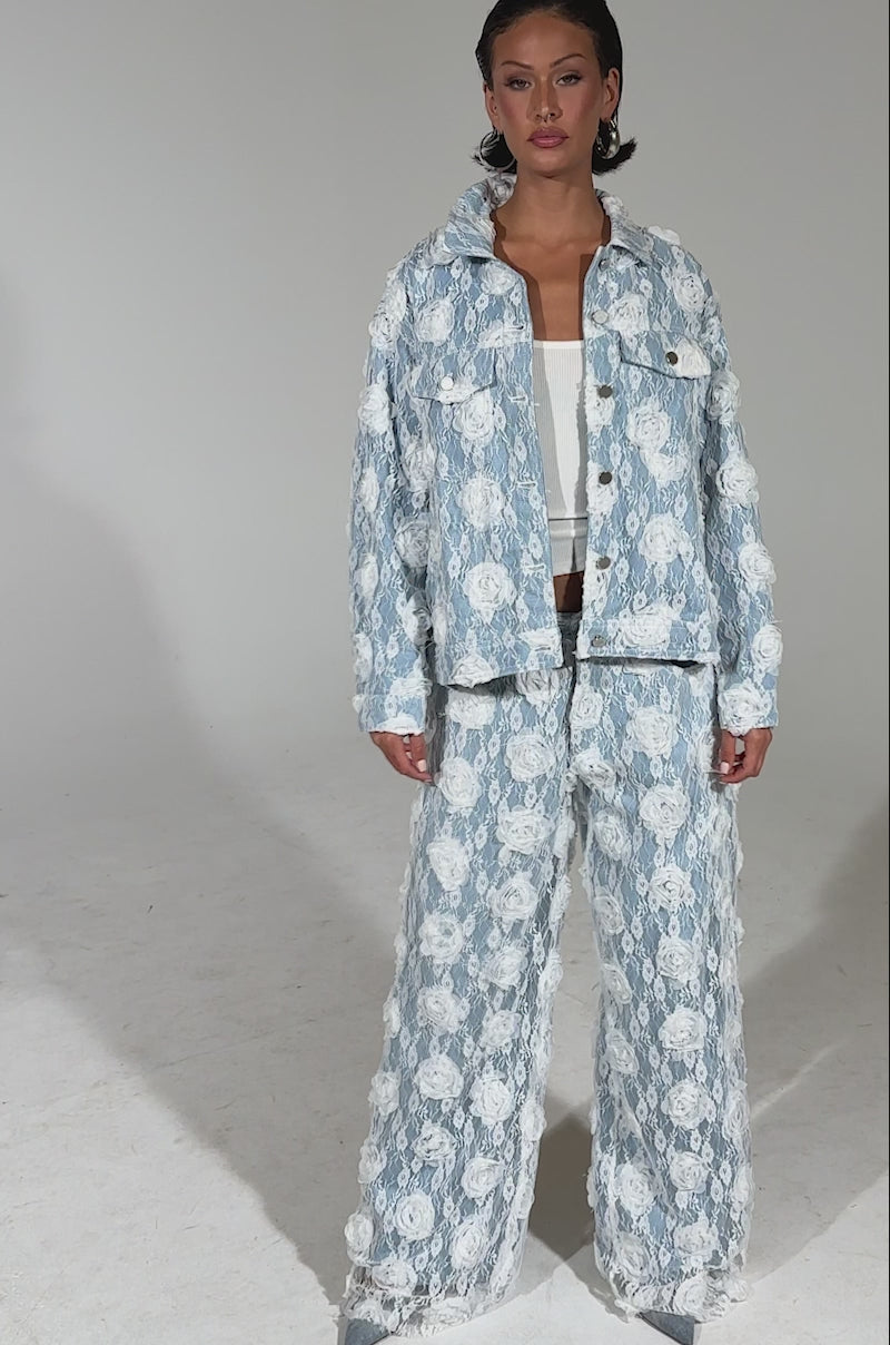 A video of a woman posing confidently against a plain background, wearing the FOLLOW MY LEAD APPLIQUE DENIM JACKET in light blue and white with a floral lace applique pattern, styled over a white top and matching pants.