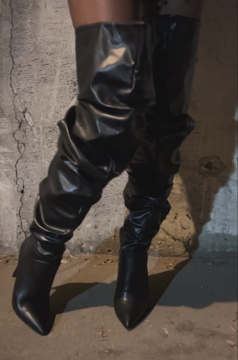 Video of woman modeling the AZALEA WANG TORVIS Ruched Stiletto Boot in Black and a black mini skirt with metallic embellishments stands on a concrete floor before a white garage door.
