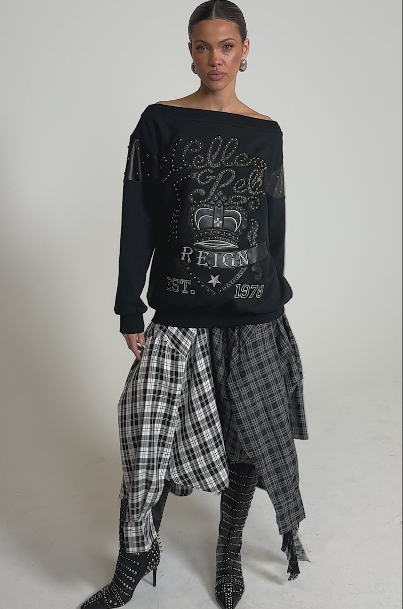 A video of a woman wearing the ROYAL OVERSIZED SWEATSHIRT with an off-the-shoulder design, paired with a layered plaid skirt and black studded heeled boots, against a simple light background.