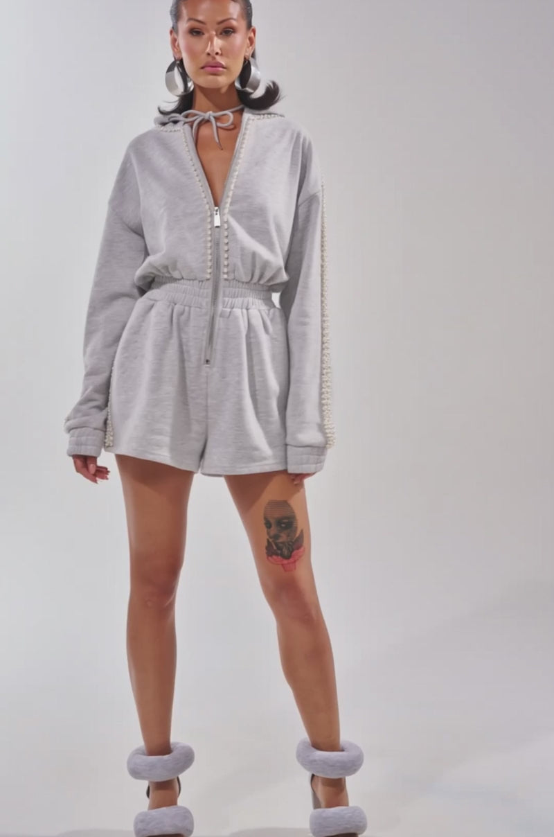 Video of woman modeling the PEARLS FOR THE GIRLS FRENCH TERRY ROMPER—a grey hooded romper with pearl-embellished sleeves—confidently posing against a plain background