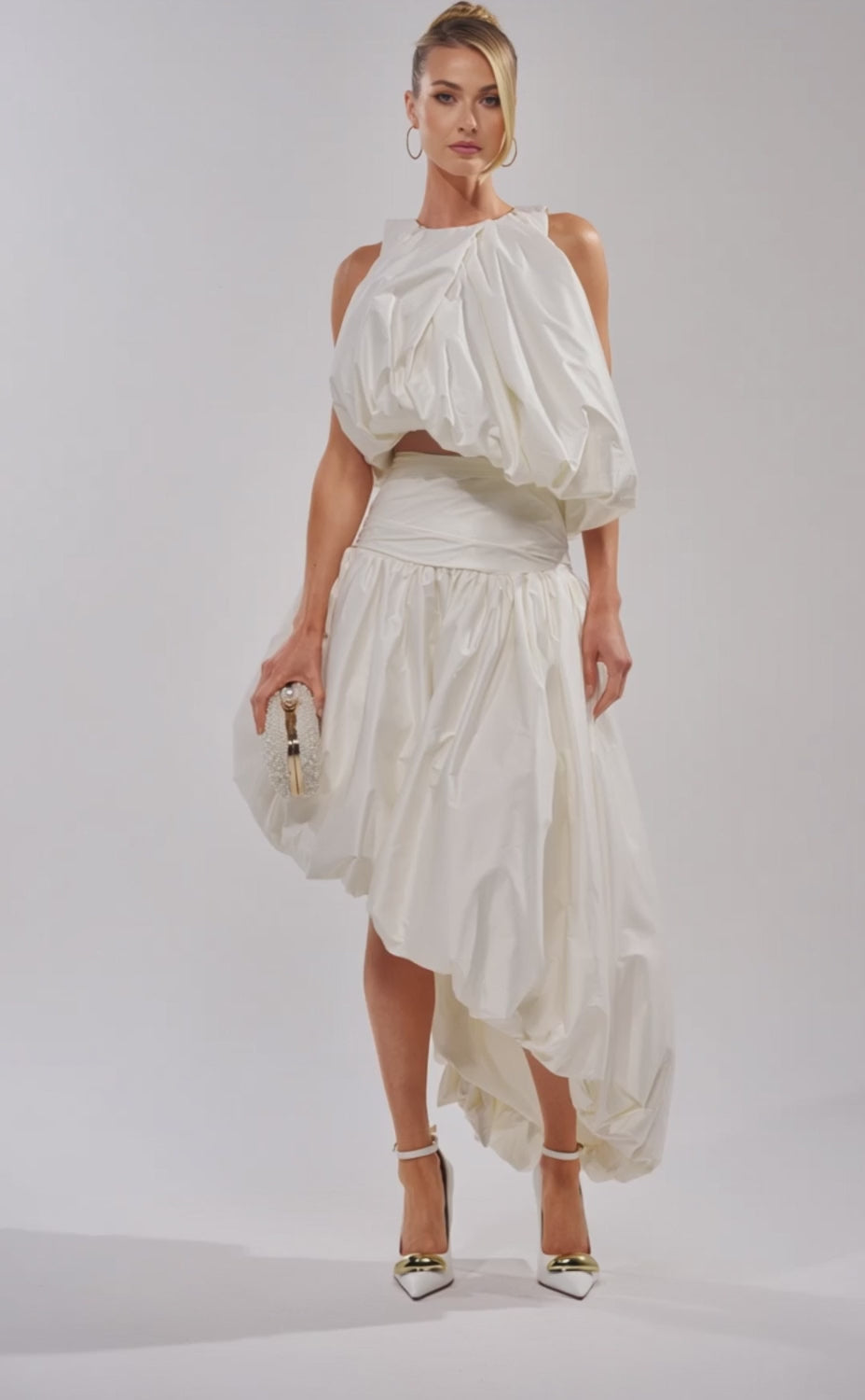 A video of a woman in the HEAD IN THE CLOUDS SLEEVELESS BLOUSE, styled with a voluminous bubble silhouette, gathered fabric, high-low hem, a textured clutch, pointed white heels, and hoop earrings.