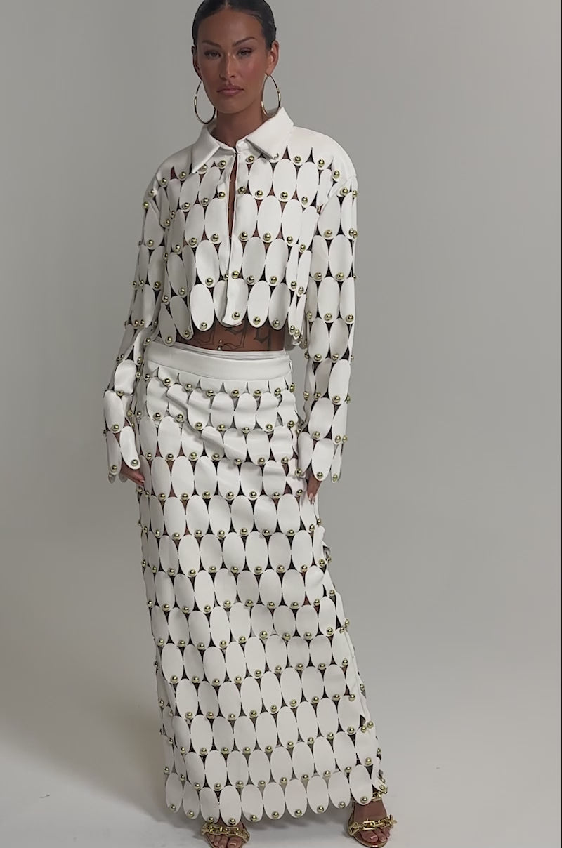 Video of woman modeling the THE SNEAK PEEK STUDDED PU MIDI SKIRT IN WHITE with a matching cream cropped jacket, gold hoop earrings, gold heels, and a small brown clutch against a plain light background.