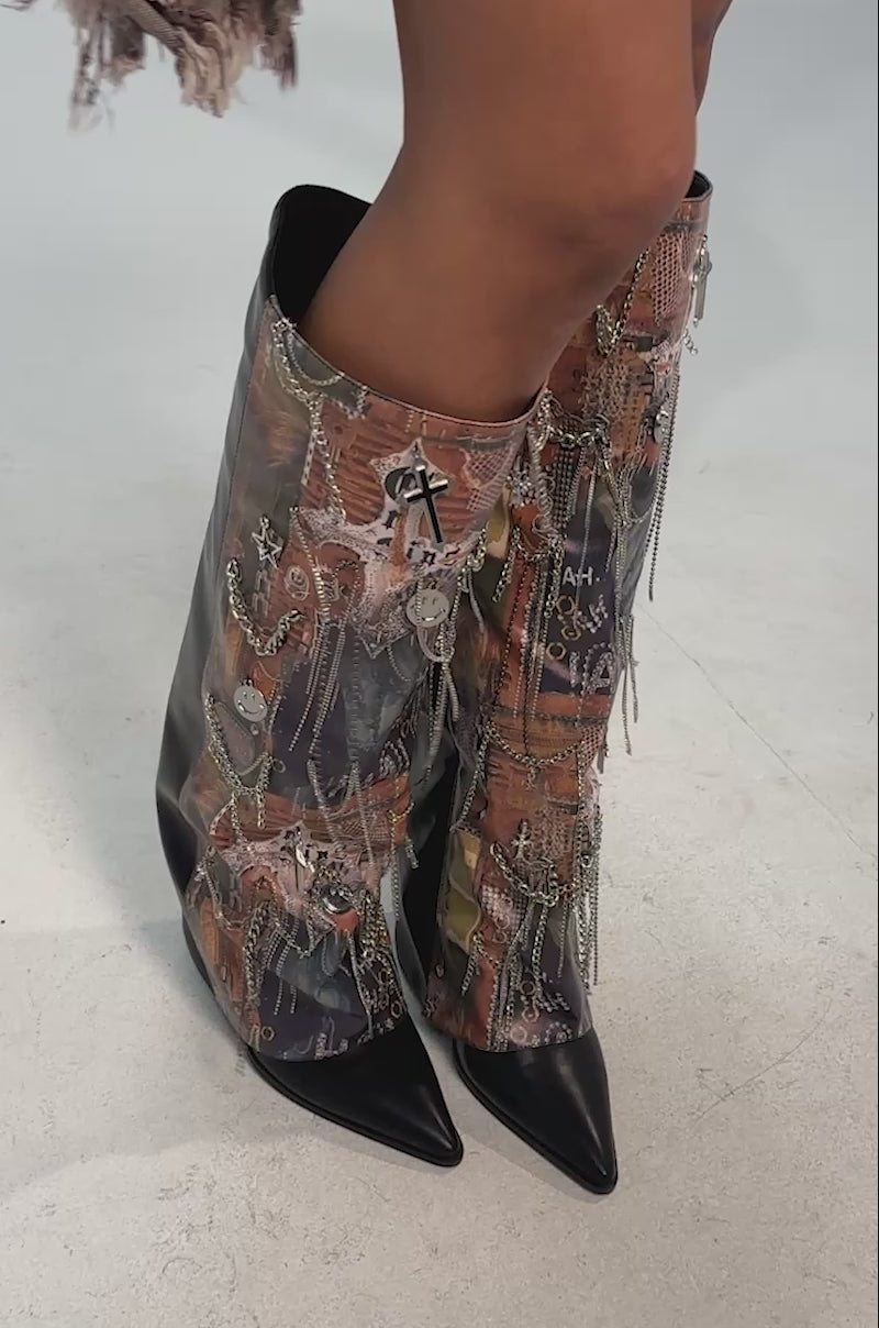 Video of woman modeling the AKIRA GOLD IVELLISE MULTI BLOCK BOOT—one side in black leather, the other with bold graphic prints, colorful patterns, embellishments, hanging chains, and pointed toes for a unique artistic look.