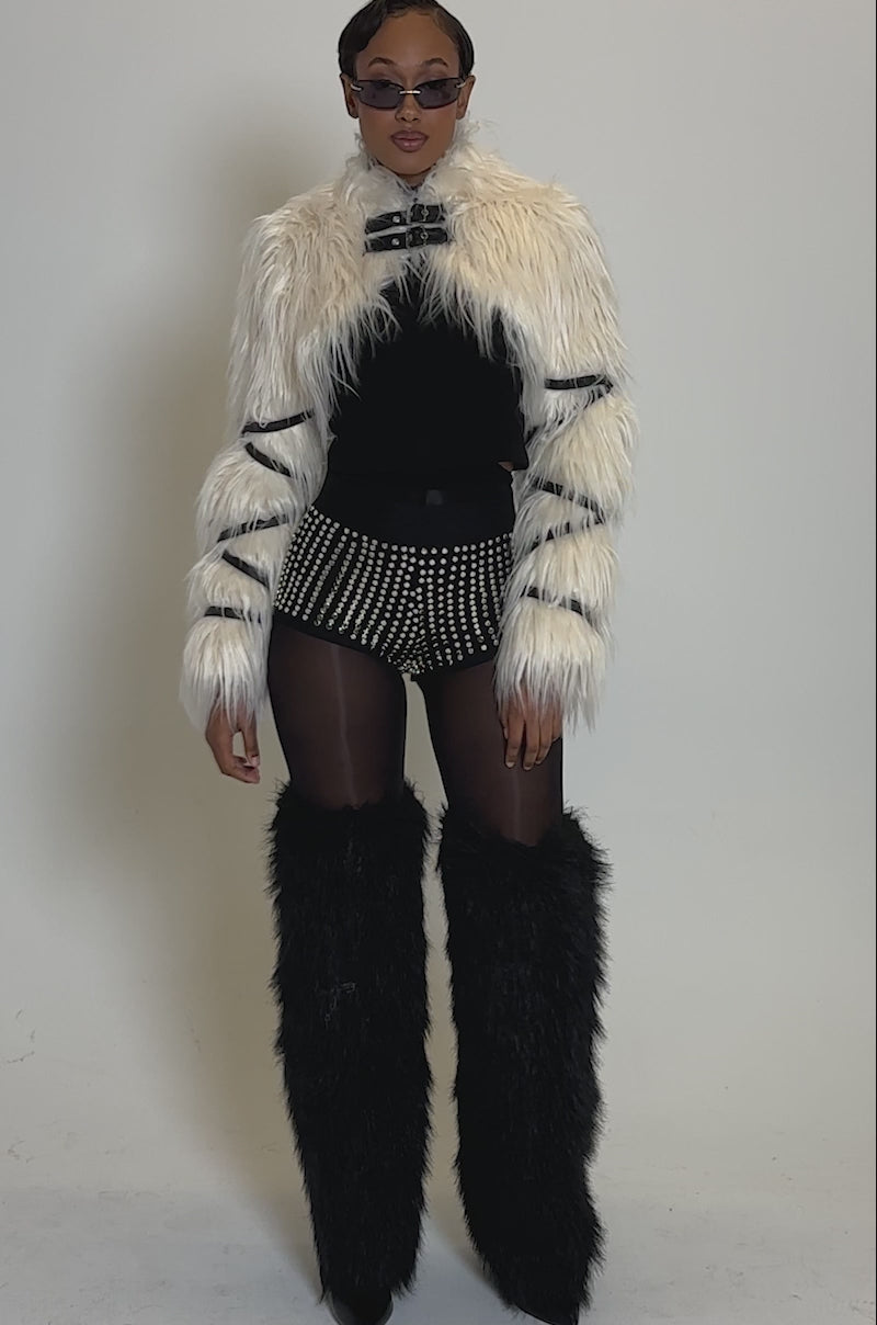 Video of woman modeling the COOLEST GIRL ON THE BLOCK MESH HOT SHORT WITH STUDS, a white faux fur cropped jacket with black straps, sheer tights, and tall black faux fur boots against a plain background.