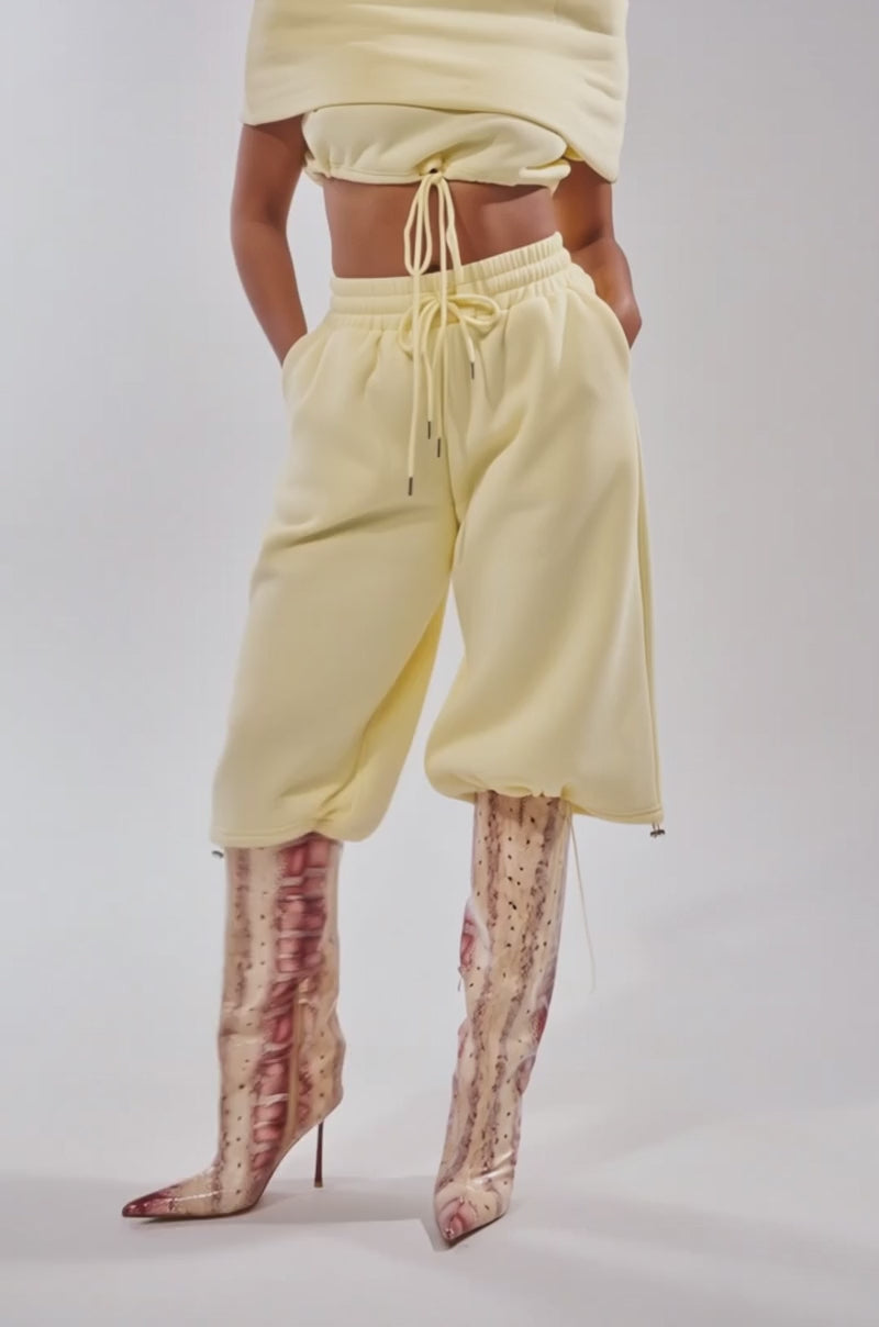 A video of a model wearing the BABY PLEASE SHORT, pale yellow jogger shorts with a high-rise elastic waistband and ankle drawstrings, tucked into tall, pointed-toe pink and beige snakeskin boots.