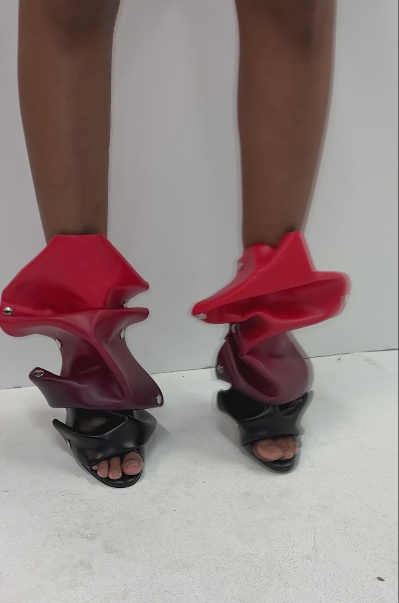 A video of a person standing against a white background, wearing short burgundy shorts and high-heeled sandals with bold, sculptural red and black ruffles at the ankles, channeling the striking style of the AZALEA WANG BIRDSONG RED OMBRE SNAP OPEN TOE BOOTIE.