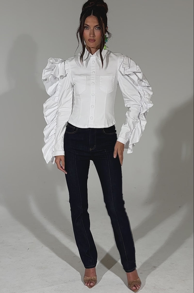 A video of a woman standing against a plain background, wearing the DREA RUFFLE SLEEVE BUTTON DOWN with dramatic sleeves, dark blue curve-hugging jeans, and open-toe heels. Her hair is styled in a loose updo.