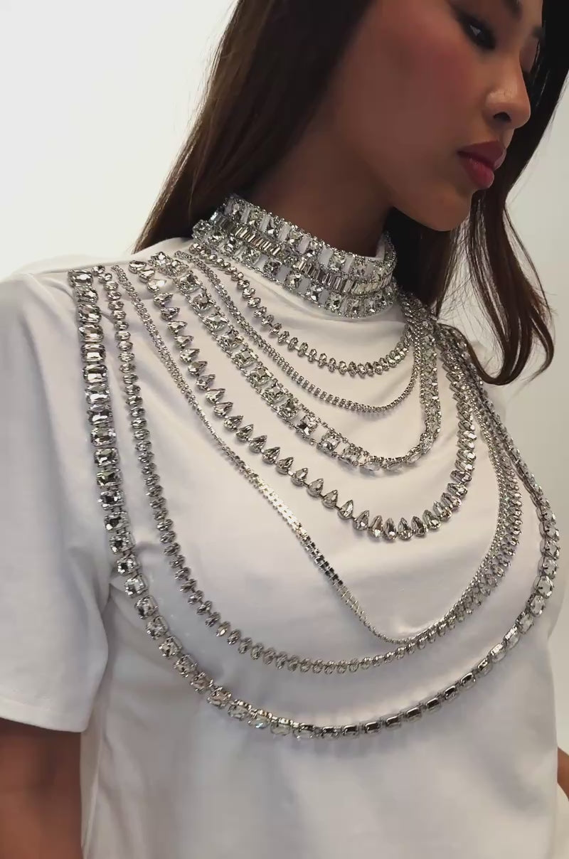 A video of a woman in a white pleated skirt and the ROYALTY RHINESTONE TEE IN WHITE, adorned with layered silver chains and holding a small clutch.