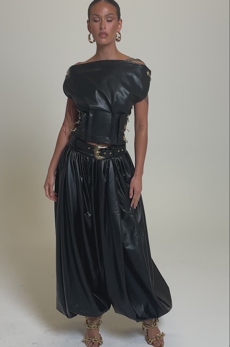 Video of woman modeling the ZONA BALLOON LEATHER TROUSERS and a black off-shoulder leather corset top, accessorized with chunky gold jewelry and chain-detailed heels, set against a plain light gray background.