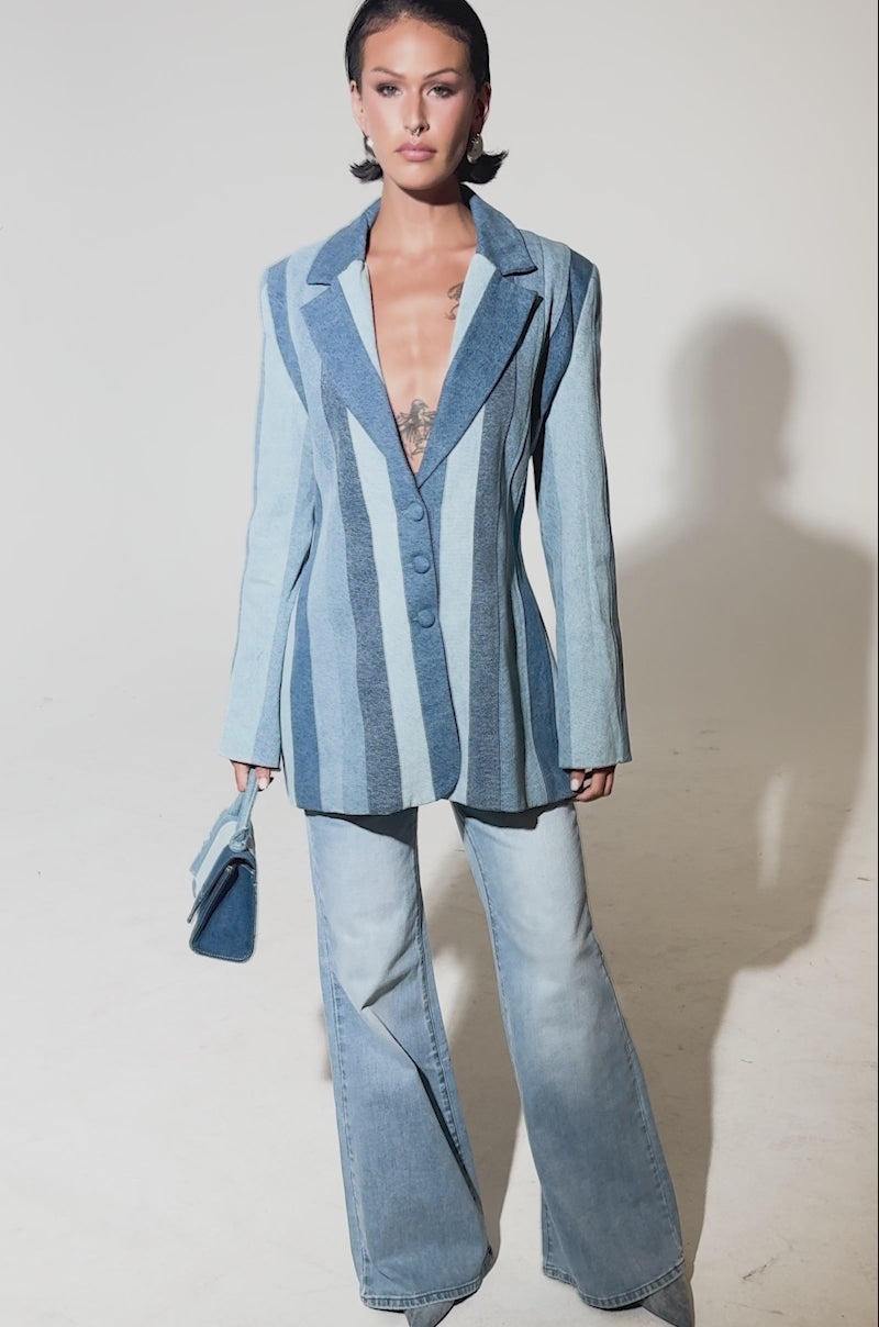 A video of a woman in the COME SLIDE PATCHWORK DENIM BLAZER, paired with flared light blue jeans and a small blue handbag. She sports short, styled hair and displays visible chest tattoos.