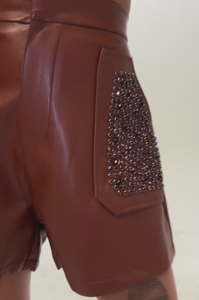 Video of a woman modeling the HOW ABOUT THIS FAUX LEATHER RHINESTONE SHORT—high-rise, brown faux leather shorts featuring large rhinestone-adorned front pockets.