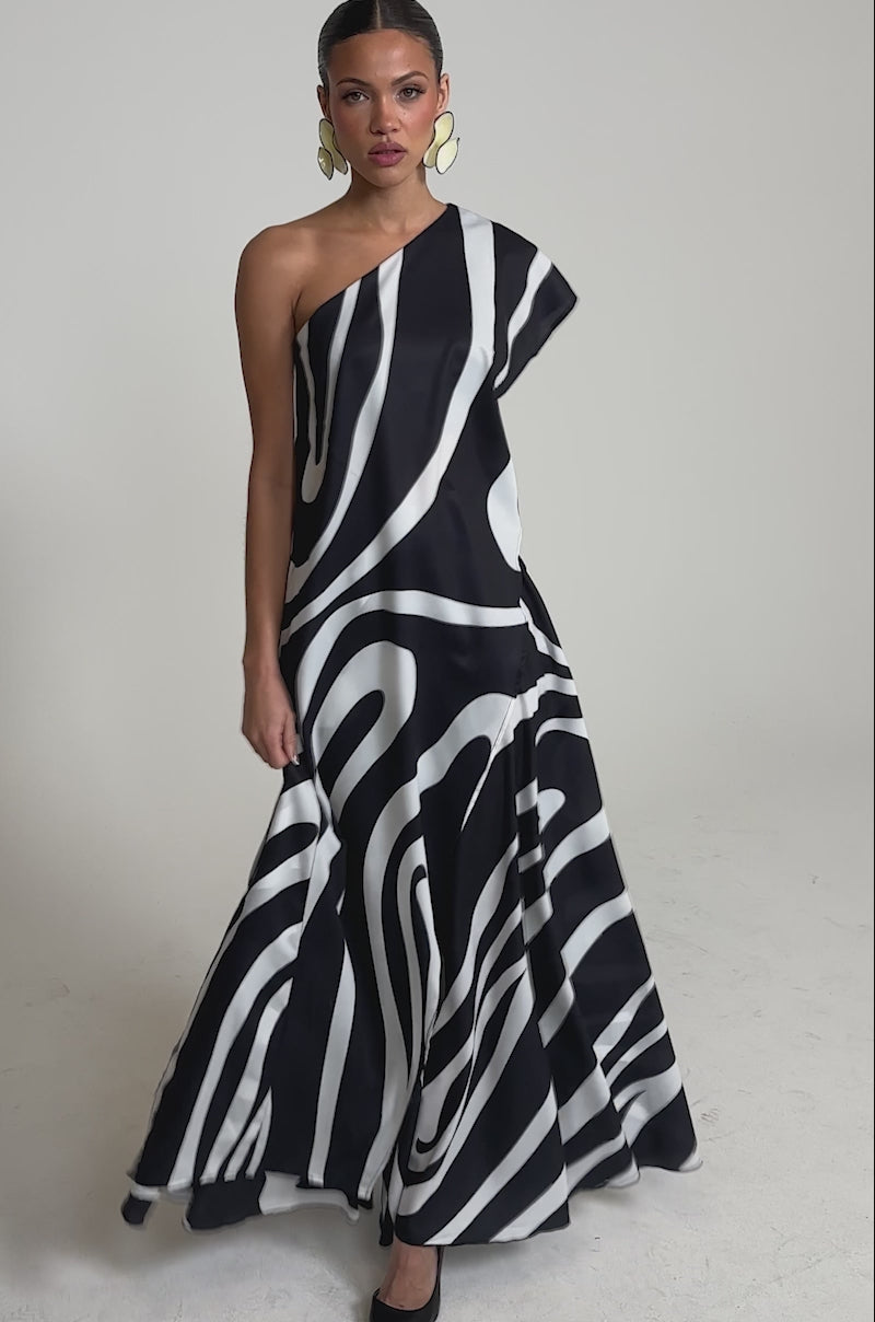 Video of woman modeling the TRINA ELONGATED SINGLE SHOULDER TOP in a bold black and white swirl print, styled with slicked-back hair, large gold hoop earrings, and black pointed-toe heels against a plain background.