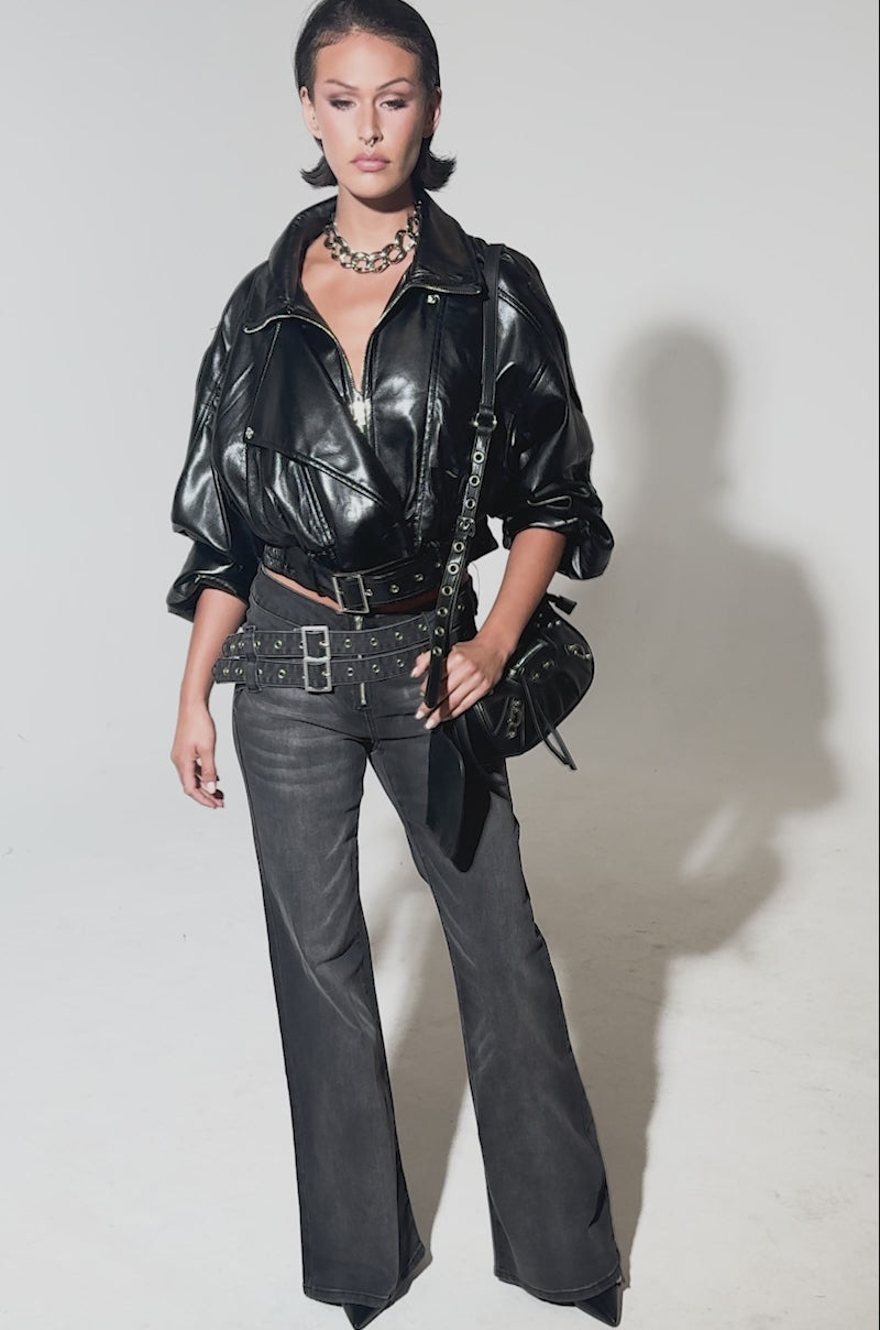 A video of a woman posing confidently in WONT LOSE MYSELF WIDE LEG DENIM jeans with an adjustable waistbelt, styled with a black leather jacket, chunky chain necklace, and black shoulder bag against a plain gray background.