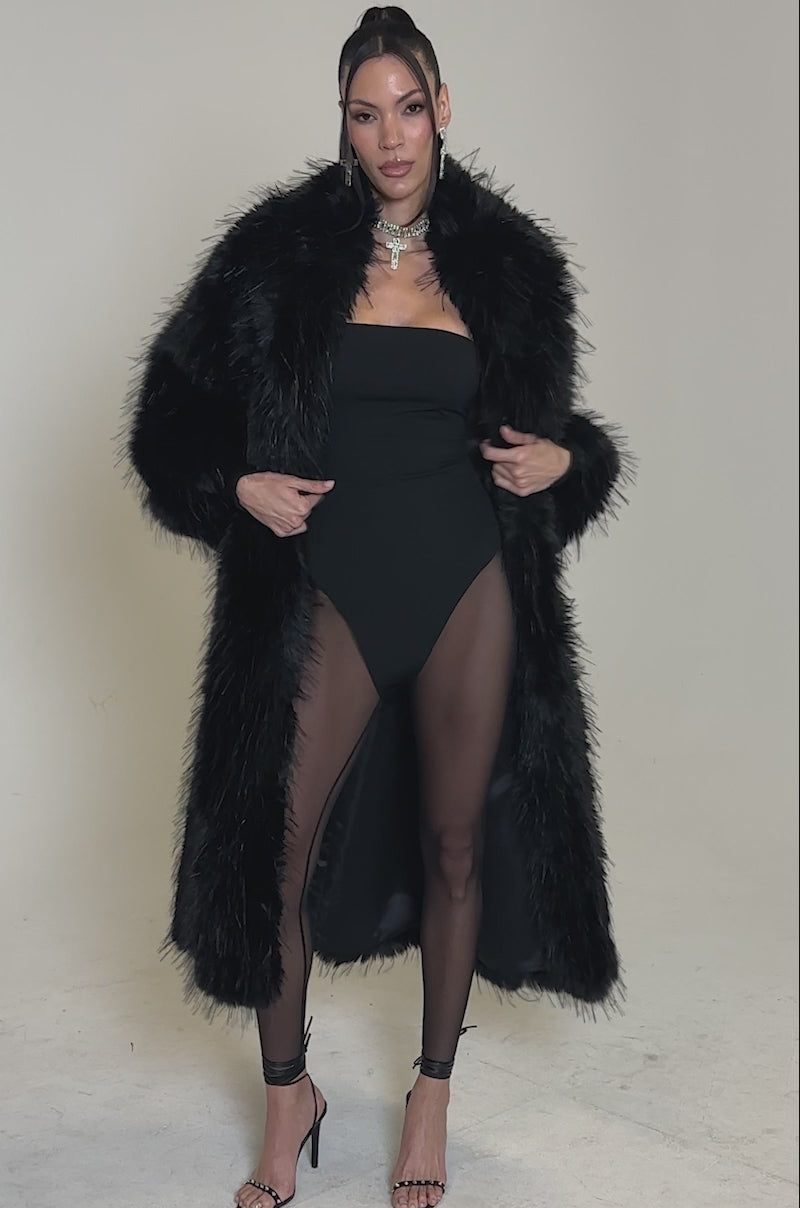 A video of a woman in the TSUNAMI FAUX FUR COAT IN BLACK, paired with a black strapless bodysuit, sheer tights, strappy heels, and layered necklaces against a plain light background.