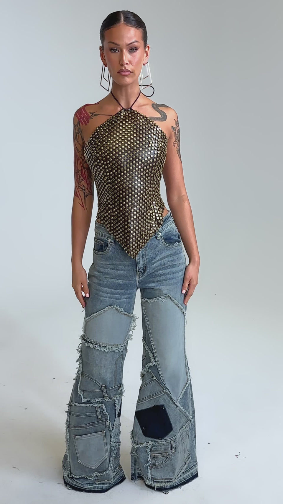 Video of woman modeling the GET FUNKY BEADED TOP IN BROWN and blue jeans. She has visible arm tattoos and large geometric earrings, standing against a plain background.