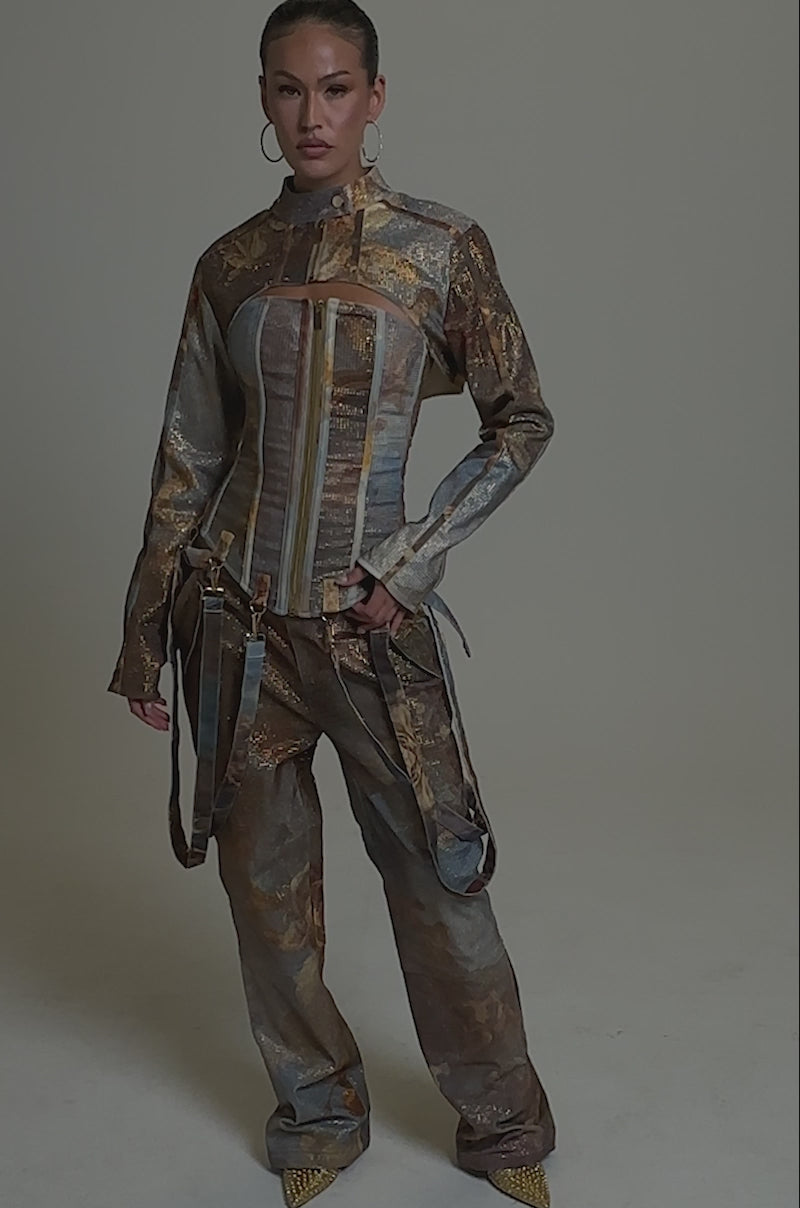 A video of a woman standing confidently in the IN THE FUTURE RHINESTONE BOLERO, a graphic print jacket with earth-toned patterns, gold accents, and decorative straps, paired with matching wide-leg pants against a plain gray background.