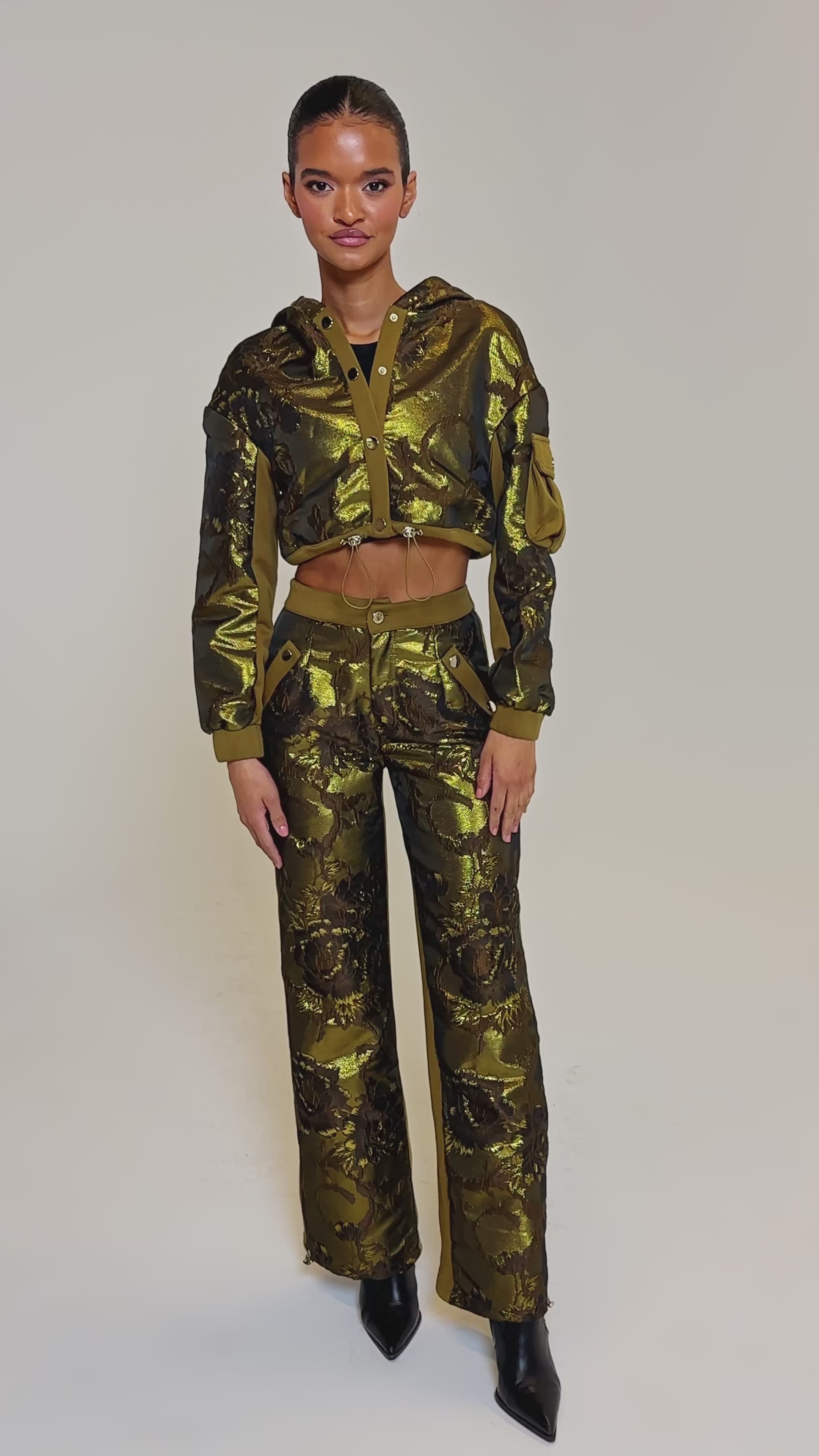 Video of woman modeling the YOU KNOW IT BABY CROP BUTTON DOWN—an olive green and gold floral-patterned AKIRA Label crop jacket with a hood and matching high-waisted pants—standing against a plain light background.