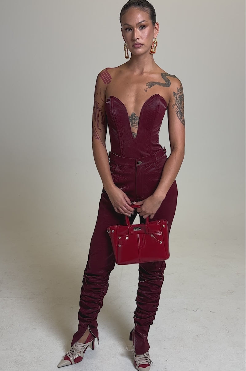 A video of a woman posing against a plain background in a strapless, deep red velvet jumpsuit with a plunging neckline and ruched legs. She accessorizes with gold earrings, pointed heels, a red handbag, and the TOO HOT FOR YOU FAUX LEATHER CORSET for extra edge.