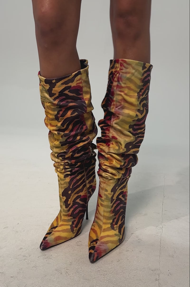 A video of a model wearing the AZALEA WANG SERIOUSLY TIGER SLOUCHY STILETTO BOOT. The boot is a knee-high, pointed-toe faux suede boot with a ruched shaft and a bold yellow, orange, and black tiger stripe print.