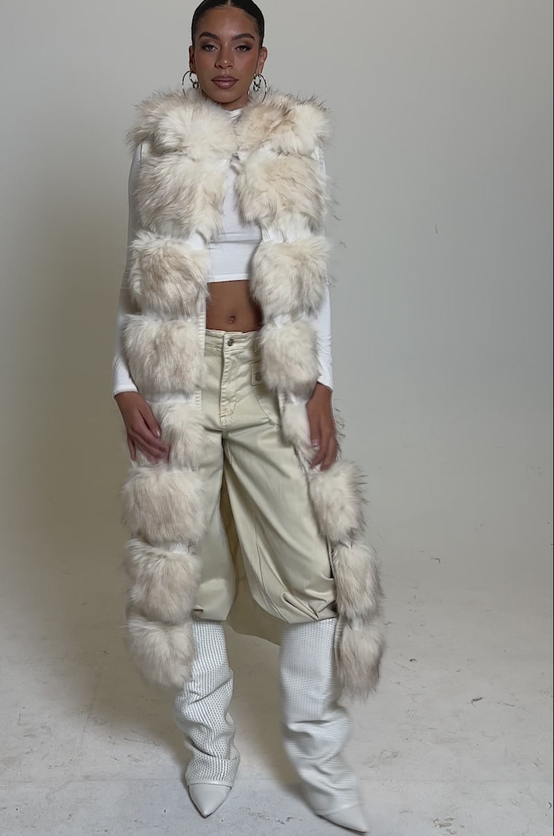 A video of a woman in the LUXURIOUS FAUX FUR HOODED VEST over a white crop top and beige high-waisted pants, paired with white heeled boots and large hoop earrings, standing against a plain background.