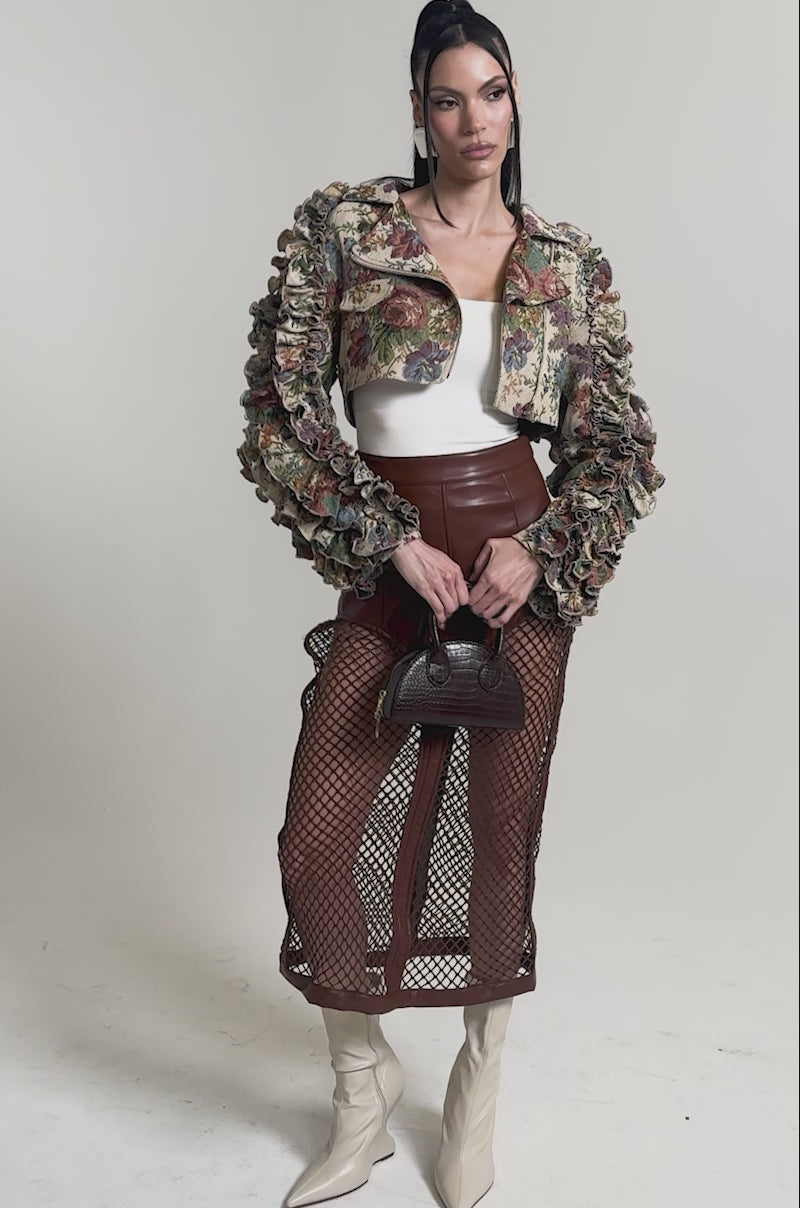 A video of a woman in the MAKE IT LOOK EASY TAPESTRY MOTO jacket with ruffled details, paired with a white top, brown leather skirt with fishnet panels, cream boots, small brown handbag, sleek straight hair, and statement earrings.