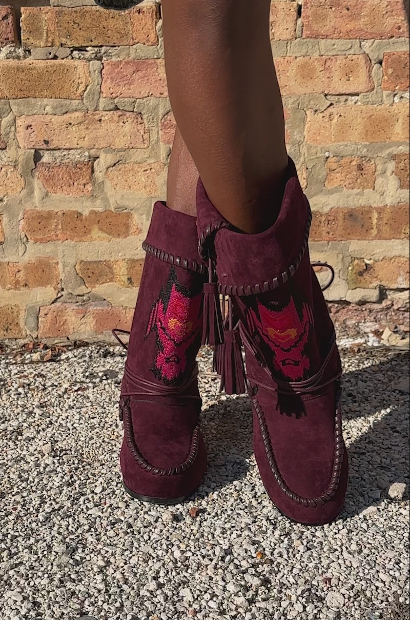 A video of a model standing on gravel before a rustic brick wall, wearing distressed denim shorts and the AZALEA WANG WERET PLUM FLAT BOOTIE, featuring fringe and tribal designs.