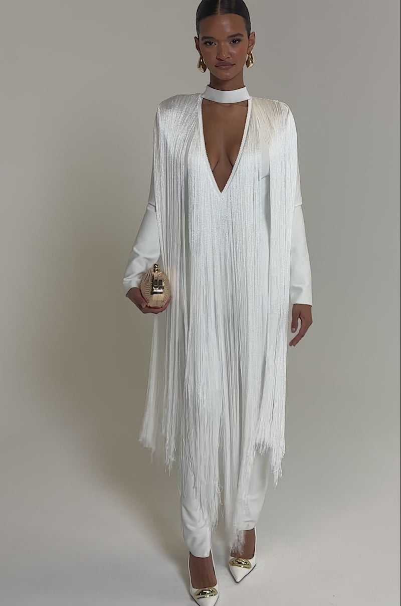 Video of woman modeling the BELLE BANDAGE JUMPSUIT WITH FRINGE IN WHITE, featuring a deep V-neck and matching choker. She pairs it with gold heels, earrings, a gold clutch, and sleeked-back hair for a chic look.