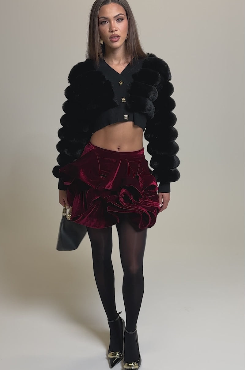 Video of woman modeling the  WILD HEART FAUX FUR CARDIGAN IN BLACK with a deep red ruffled velvet mini skirt, holding a black handbag with a gold handle. She has straight, shoulder-length hair and a neutral expression.