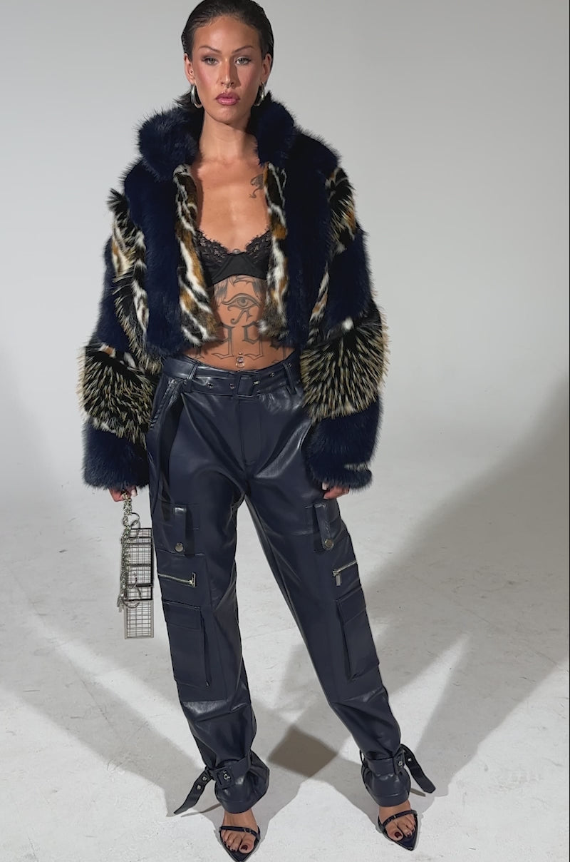 A video of a woman wearing a blue and black fur jacket over a black lace bralette, navy BUTTER LUXE PANTS, black heels, and holding a small metallic rectangular handbag stands confidently against a plain background.