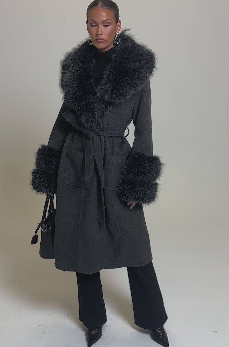 Video of woman modeling the HARMONY FUR TRIM TRENCH, a long dark gray winter coat with large faux fur trim on the collar and cuffs, holding a black handbag against a plain light gray background.