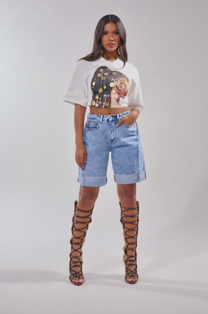 A video of a woman in the LOCKED AND LOADED EMBELLISHED TEE, paired with high-waisted cuffed denim shorts and knee-high brown lace-up gladiator sandals.
