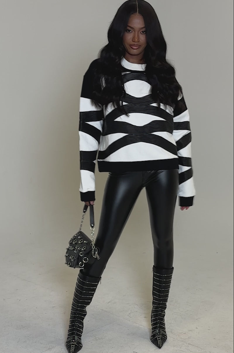 A video of a woman with long wavy hair wearing the bold CROSSING LINES SWEATSHIRT, shiny black leather leggings, studded knee-high boots, and carries a small handbag against a plain light gray backdrop for standout streetwear style.