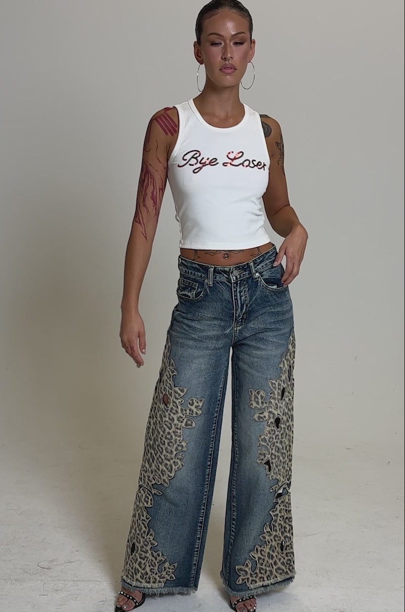 A video of a woman standing confidently in the LOSER BYE TANK, a ribbed knit top with bold red text, paired with wide-leg blue jeans with lace cutouts, and sandals.